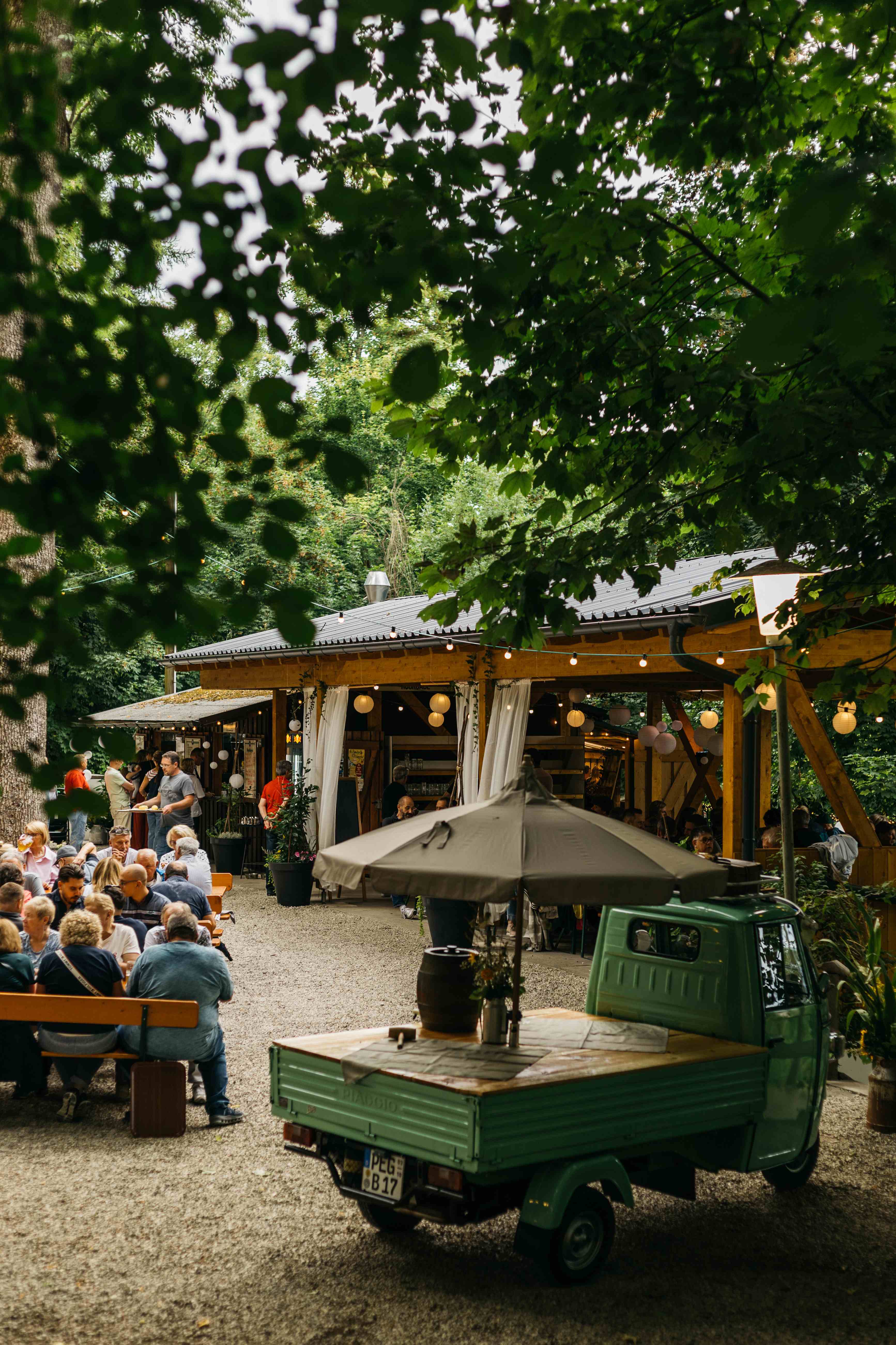 Gut besuchter Biergarten am Schlossberg unter Bäumen, mit Lichterketten und überdachtem Bereich